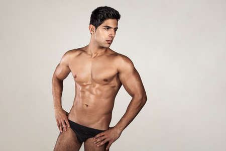 Portrait of handsome man with tanned athletic body, demonstrating his perfect sporty body, looking away, keeps hands on hips, wearing black panties. Indoor studio shot isolated on gray background.の写真素材