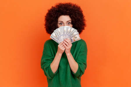 Portrait of woman with Afro hairstyle wearing green casual style sweater covering face with dollars...