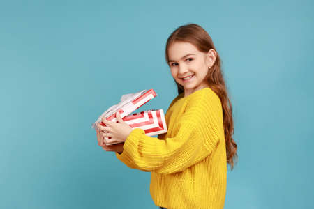 Side view of little girl opens gift box, smiling to camera, celebrating birthday, Christmas holiday, wearing yellow casual style sweater. Indoor studio shot isolated on blue background.