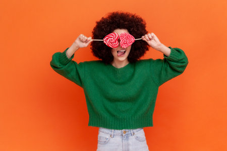 Playful childish woman with Afro hairstyle wearing green casual style sweater covering heart shape lollipops, hiding, having fun with sweets. Indoor studio shot isolated on orange background.の写真素材