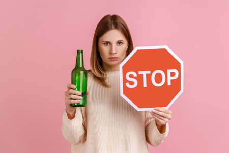 Portrait of serious blond woman holding red stop sign and bottle with alcoholic beverage, calls on dont drink alcohol, wearing white sweater. Indoor studio shot isolated on pink background.の写真素材