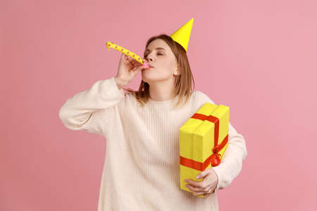 Portrait of happy blond woman in party cone, holding wrapped present box, blowing horn, celebrating...