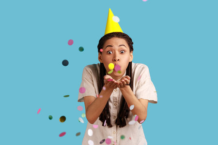 Portrait of excited woman with black dreadlocks and party cone, blowing confetti, celebrating birthday, festive mood, wearing white shirt. Indoor studio shot isolated on blue background.の写真素材