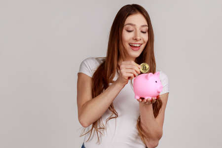 Portrait of happy smiling dark haired woman investing in bitcoins, putting golden crypto coin into...