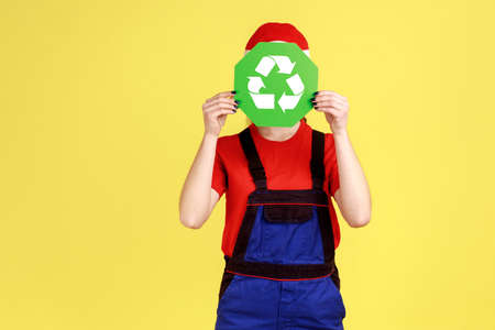 Portrait of unknown anonymous worker woman standing hiding her face behind green recycling sign, ecology, wearing overalls and red cap. Indoor studio shot isolated on yellow background.の写真素材