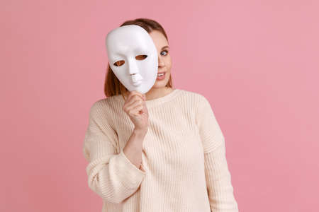 Portrait of smiling blond woman holding white mask, peeking, looking at camera, pretending to be another person, wearing white sweater. Indoor studio shot isolated on pink background.の写真素材