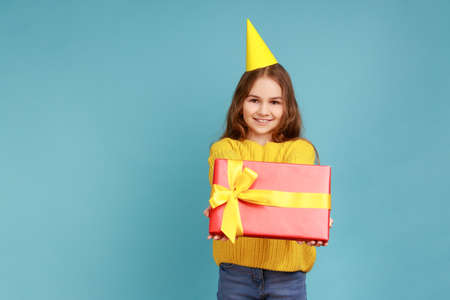 Portrait of little girl in party cone giving present box, congratulating her friend with birthday, wearing yellow casual style sweater. Indoor studio shot isolated on blue background.の写真素材