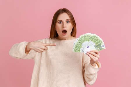 Portrait of amazed shocked blond woman holding pointing at many euro banknotes looking at camera,...