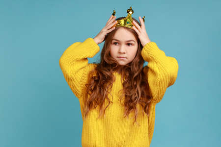 Portrait of little princess girl standing and wear golden diadem crown with smile, looking at camera, wearing yellow casual style sweater. Indoor studio shot isolated on blue background.の写真素材