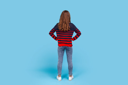 Full length back portrait of woman wearing striped casual style sweater, posing backwards with hands on hips, standing calm unrecognizable, waiting. Indoor studio shot isolated on blue background.の写真素材