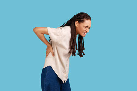 Woman with black dreadlocks has backache, suffers from painful muscle injury, smirks face from unpleasant feelings, wearing white shirt. Indoor studio shot isolated on blue background.の写真素材