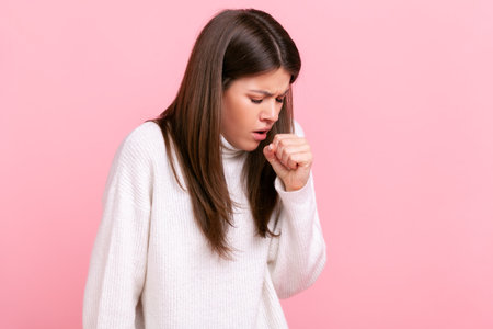 Profile portrait of young adult brunette sick woman coughing, catches cold, having high temperature, wearing white casual style sweater. Indoor studio shot isolated on pink background.の写真素材