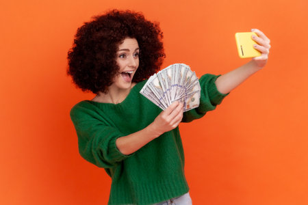 Excited woman with Afro hairstyle wearing green casual style sweater making selfie with big fan of dollar banknotes, showing money to followers. Indoor studio shot isolated on orange background.の写真素材