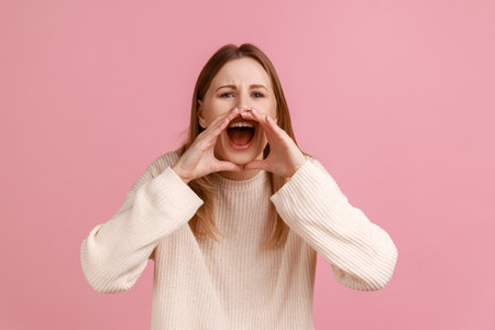 Portrait of aggressive young adult blond woman standing, holding arms near wide open mouth and screaming loud, wearing white sweater. Indoor studio shot isolated on pink background.の写真素材
