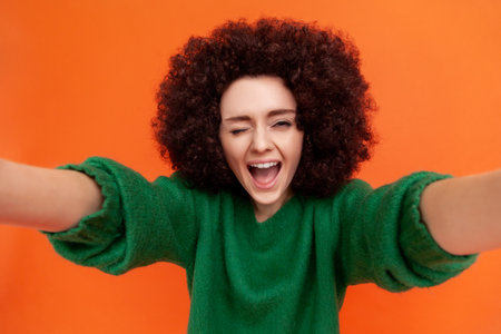 Playful woman with Afro hairstyle wearing green casual style sweater making selfie or broadcasting livestream POV, winking and keeping mouth open. Indoor studio shot isolated on orange background.の写真素材
