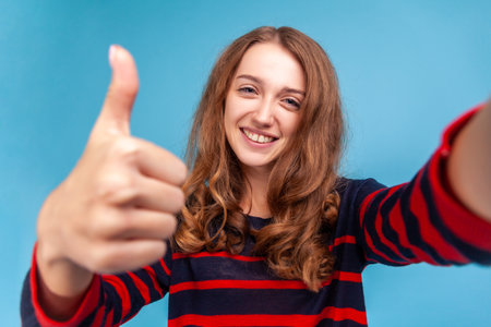 Portrait of positive woman wearing striped casual style sweater, being in good mood, showing thumb down like gesture, taking selfie POV. Indoor studio shot isolated on blue background.の写真素材