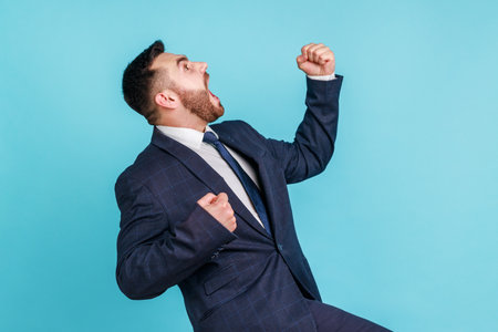 Side view of overjoyed handsome man wearing official style suit expressing winning gesture with raised fists and screaming, celebrating victory. Indoor studio shot isolated on blue background.