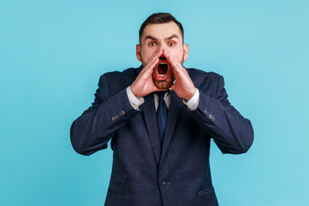 Portrait of hysterical young adult man wearing official style suit loudly yelling in panic, holding hands near his mouth, teenager protest. Indoor studio shot isolated on blue background.の写真素材