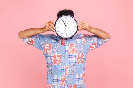 Unknown man in blue casual style shirt hiding face behind big wall clock display, wasting his time, procrastination, bad organization of working time. Indoor studio shot isolated on pink background.の写真素材