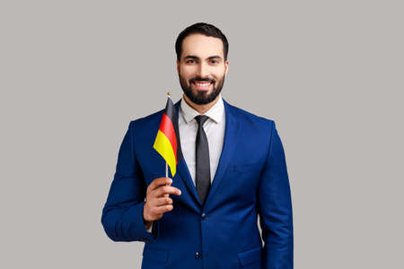 Delighted businessman holding Germany flag, celebrating Day of Germany - 3th October, expressing positive emotions, wearing official style suit. Indoor studio shot isolated on gray background.の写真素材