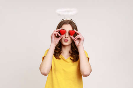 Romantic young female halo covering eyes with toy hearts as if looking with love, making kissing gesture, affection adoration in relations. Indoor studio shot isolated on gray background.の写真素材