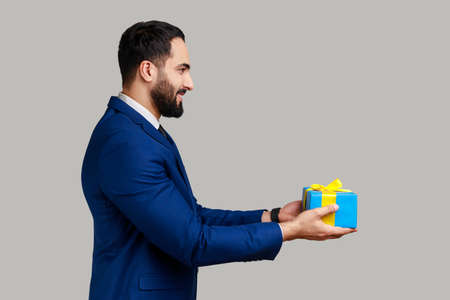 Side view portrait of smiling positive man giving blue wrapped present box, congratulating with holidays, holding gift, wearing official style suit. Indoor studio shot isolated on gray background.の写真素材