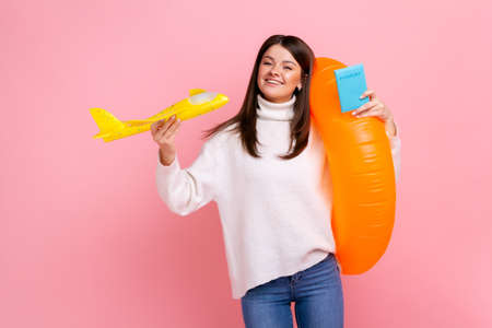 Portrait of joyful dark haired girl holding orange rubber ring, passport document and paper airplane, wearing white casual style sweater. Indoor studio shot isolated on pink background.の写真素材