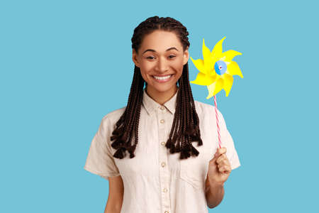 Origami hand mill. Portrait of happy smiling carefree woman with black dreadlocks holding paper windmill, pinwheel toy on stick, wearing white shirt. Indoor studio shot isolated on blue background.の写真素材