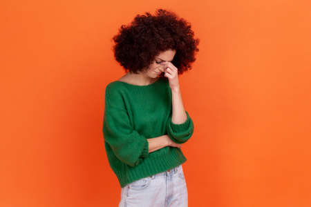 Portrait of sad woman with Afro hairstyle wearing green casual style sweater hiding face in hand crying, feeling stressed worried, Indoor studio shot isolated on orange background.の写真素材