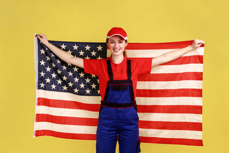 Portrait of happy smiling worker woman standing with american flag, celebrating national holiday, looking at camera, wearing overalls and red cap. Indoor studio shot isolated on yellow background.の写真素材