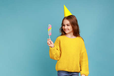 Portrait of cute little girl in party cone standing with delicious ice cream, expressing happiness, wearing yellow casual style sweater. Indoor studio shot isolated on blue background.の写真素材
