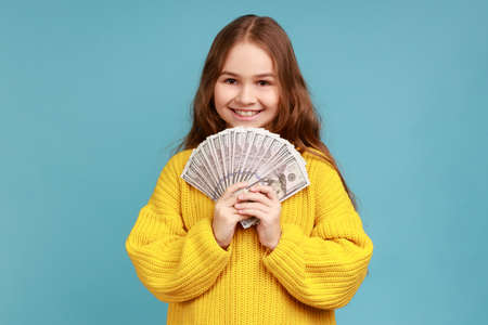 Little girl holding fan of dollar banknotes, showing big money, looking at camera with toothy smile, wearing yellow casual style sweater. Indoor studio shot isolated on blue background.の写真素材