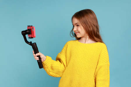 Portrait of little charming girl blogger broadcasting livestream, using steadicam and mobile phone, wearing yellow casual style sweater. Indoor studio shot isolated on blue background.の写真素材