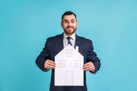 Man wearing official style suit holding paper house with drawn windows and doors, making project of home of her dreams, looking at camera with smile. Indoor studio shot isolated on blue background.の写真素材