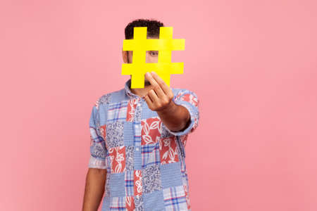 Funny unknown man blogger in blue casual style shirt hiding face behind yellow hashtag sign, calls...