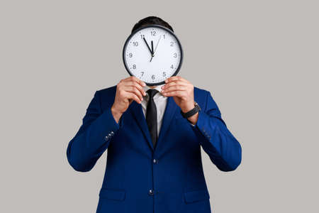 Unknown anonymous man hiding face behind big wall clock, afraid of deadline, wasting time, being late, wearing official style suit. Indoor studio shot isolated on gray background.の写真素材