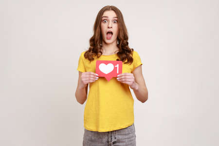 Like and follow my blog. Shocked blogger young woman in yellow T-shirt holding social media Heart button, astonishing of amount of followers. Indoor studio shot isolated on gray background.の写真素材