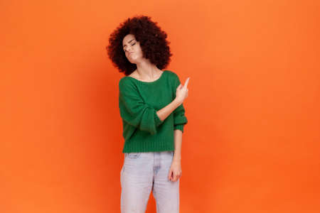 Get out. Woman with Afro hairstyle wearing green casual style sweater pointing away, scolding for bad result and showing exit, ordering to leave. Indoor studio shot isolated on orange background.の写真素材