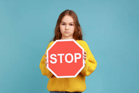 Little girl holding Stop symbol, showing red traffic sign, warning you about road safety rules, wearing yellow casual style sweater. Indoor studio shot isolated on blue background.の写真素材