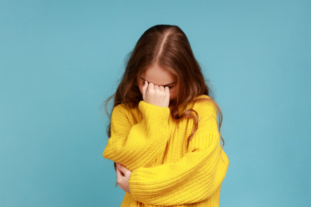 Portrait of little girl hiding face down and crying, upset about loss, feeling sorrow and regret, wearing yellow casual style sweater. Indoor studio shot isolated on blue background.の写真素材