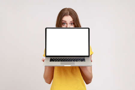 Positive unknown female in yellow T-shirt hiding half of face behind laptop with white empty display, looking at camera with big surprised eyes. Indoor studio shot isolated on gray background.の写真素材