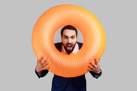 Bearded man holding rubber ring over her neck and looking at camera with surprised expression and opened mouth, wearing official style suit. Indoor studio shot isolated on gray background.の写真素材