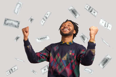 Money rain, winner and rich. Hurray. satisfied happy man with dreadlocks rising hands up with toothy smile on face, pleased, amazed with money falling. Indoor studio shot isolated on gray backgroundの写真素材