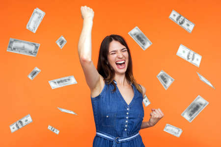Money rain, winner and rich. Hurray. satisfied happy young woman rising hands up with toothy smile on face, pleased, amazed with money falling. Indoor studio shot isolated on orange backgroundの写真素材