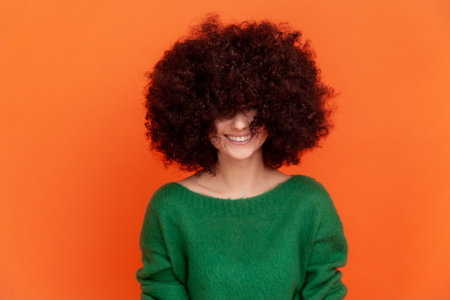 Happy positive woman with Afro hairstyle wearing green casual style sweater sowing her fluffy hair, heed hairdresser, posing with toothy smile. Indoor studio shot isolated on orange background.の写真素材