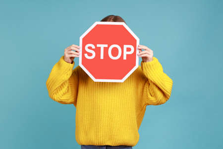 Portrait of unknown little kid covers face with Stop symbol, anonymous child holds red traffic sign, wearing yellow casual style sweater. Indoor studio shot isolated on blue background.の写真素材