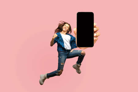 Happy young woman flying and jumping in air and showing big mobile empty screen for copy space and advertising area. indoor studio shot isolated on pink backgroundの写真素材