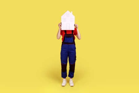 Handy woman covering face with paper house, advertising of home repair service and maintenance, help in relocation, wearing overalls and red cap. Indoor studio shot isolated on yellow background.の写真素材