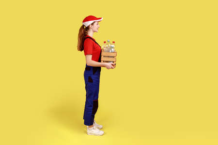 Side view portrait of positive satisfied woman volunteer standing with box full of empty plastic bottles, wearing overalls and red cap. Indoor studio shot isolated on yellow background.の写真素材