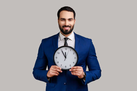 Handsome bearded businessman holding big clock and looking at camera with toothy smile, playful...